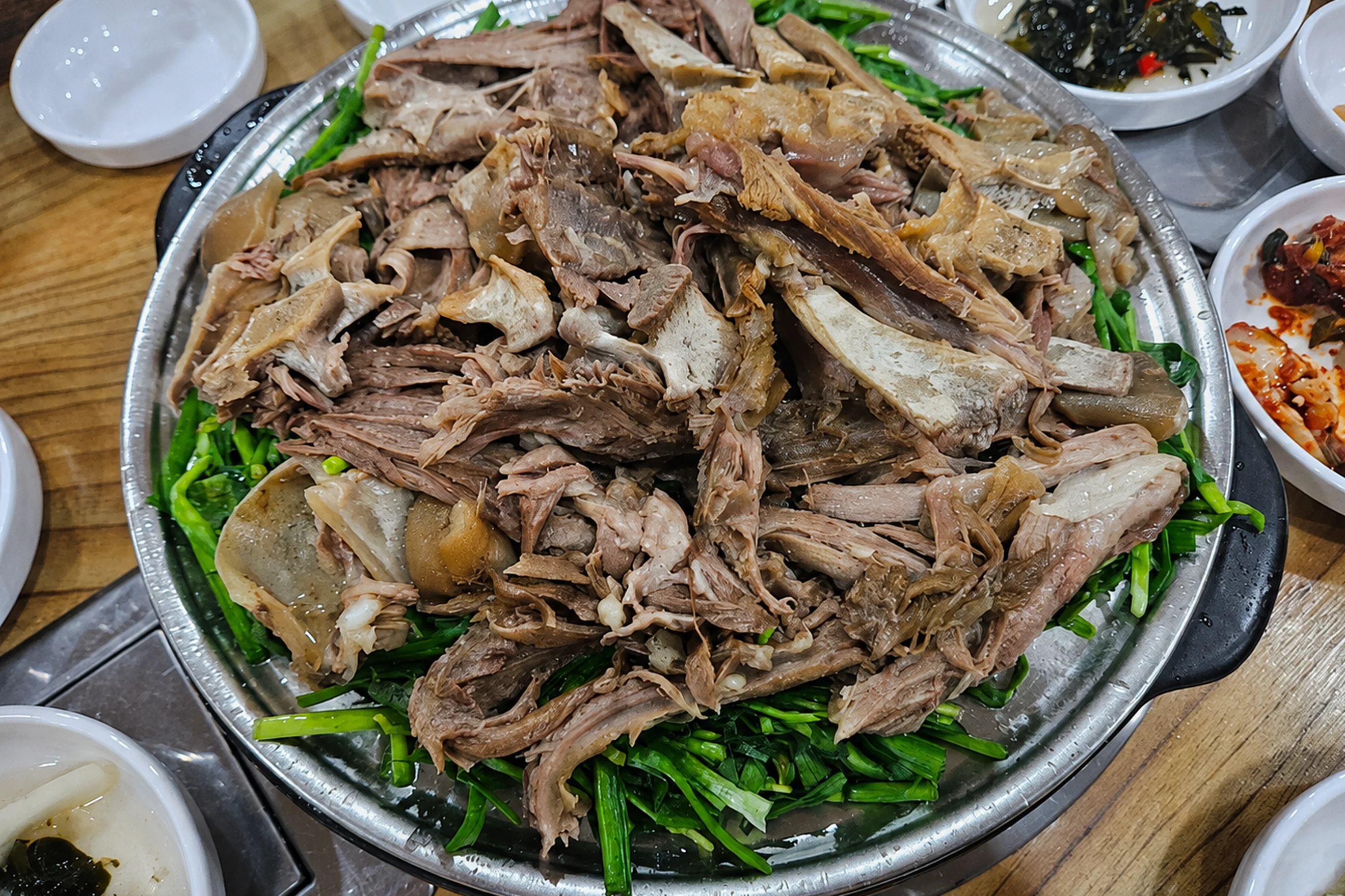 둥근 금속 그릇에 고기와 채소가 담기고 주변에 반찬이 놓인 음식 사진